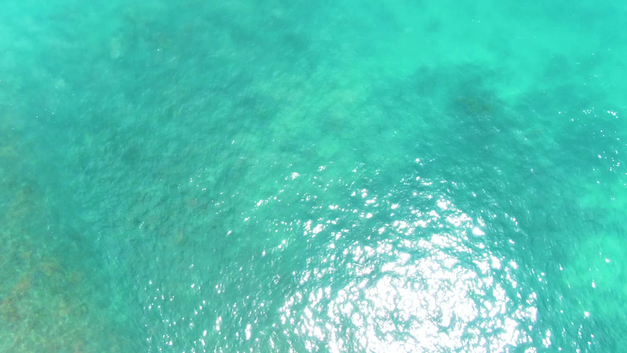Aerial frame over Punta Buzzards turquoise water with a concentrated white sun glint, soft ripples, and faint seabed color variation, no visible people.