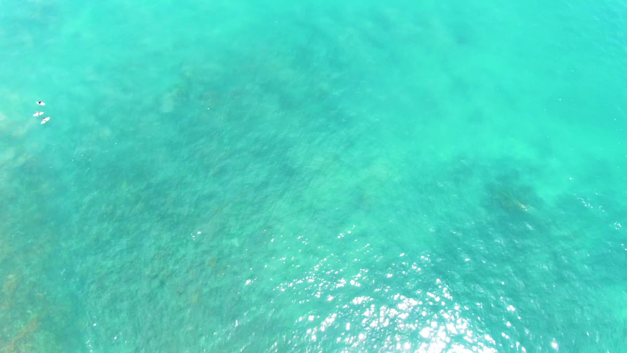 Top-down Punta Buzzards ocean view with turquoise water, a bright lower reflection area, and tiny white swimmer shapes near the upper center.