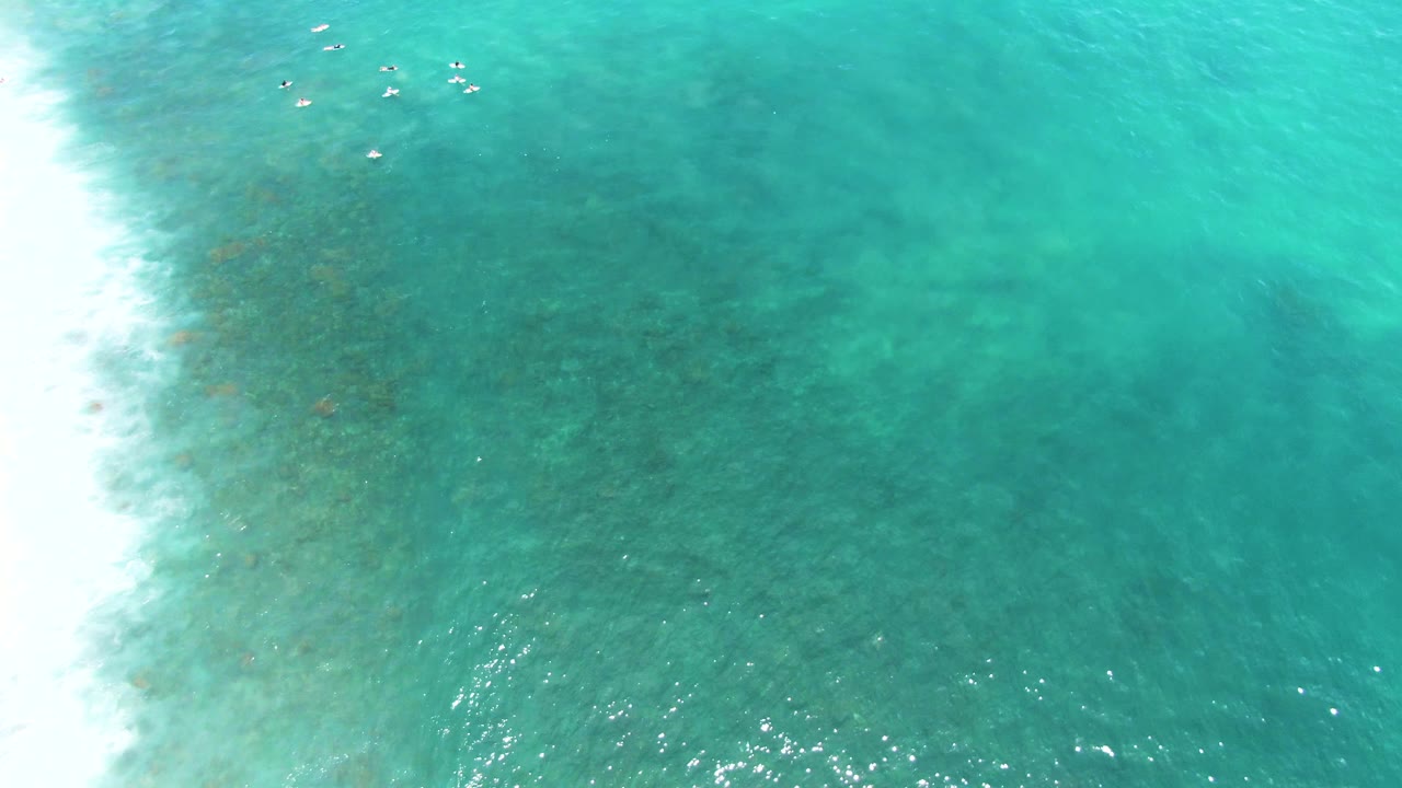 Overhead view at Punta Buzzards, Baja California Sur, with white shoreline at the left edge, turquoise shallow water, visible seabed texture, and tiny distant people near the surf.
