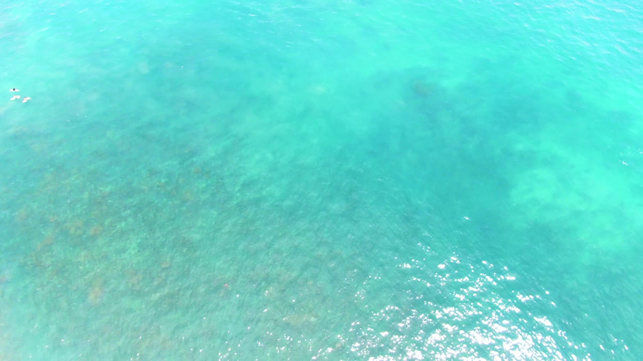 High overhead view of shallow turquoise water at Punta Buzzards, with submerged rocky or coral patches visible and tiny distant people near the upper center.