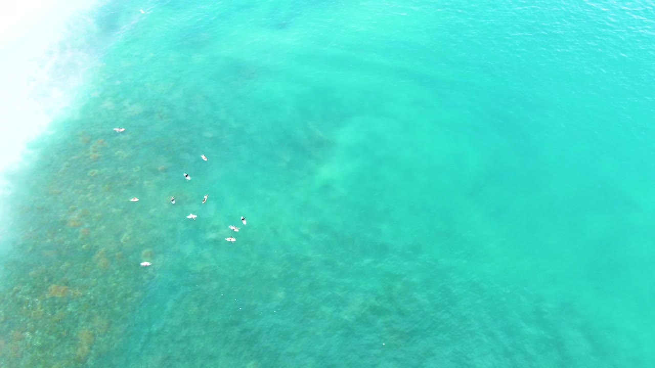 Aerial frame from Punta Buzzards showing turquoise shallow water, a white surf line at left, darker seabed patches, and a loose cluster of swimmers near the center.