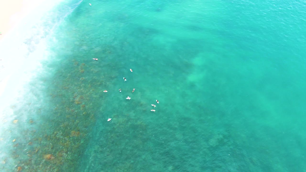 Aerial frame from Punta Buzzards where the swimmers sit right of the surf wash, with darker seabed texture visible under the turquoise water.