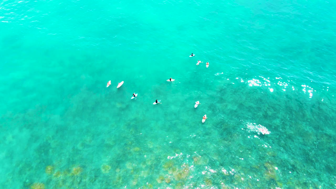 Overhead view at Punta Buzzards, Baja California Sur, with scattered people on surfboards crossing turquoise water and faint darker seabed patches below.