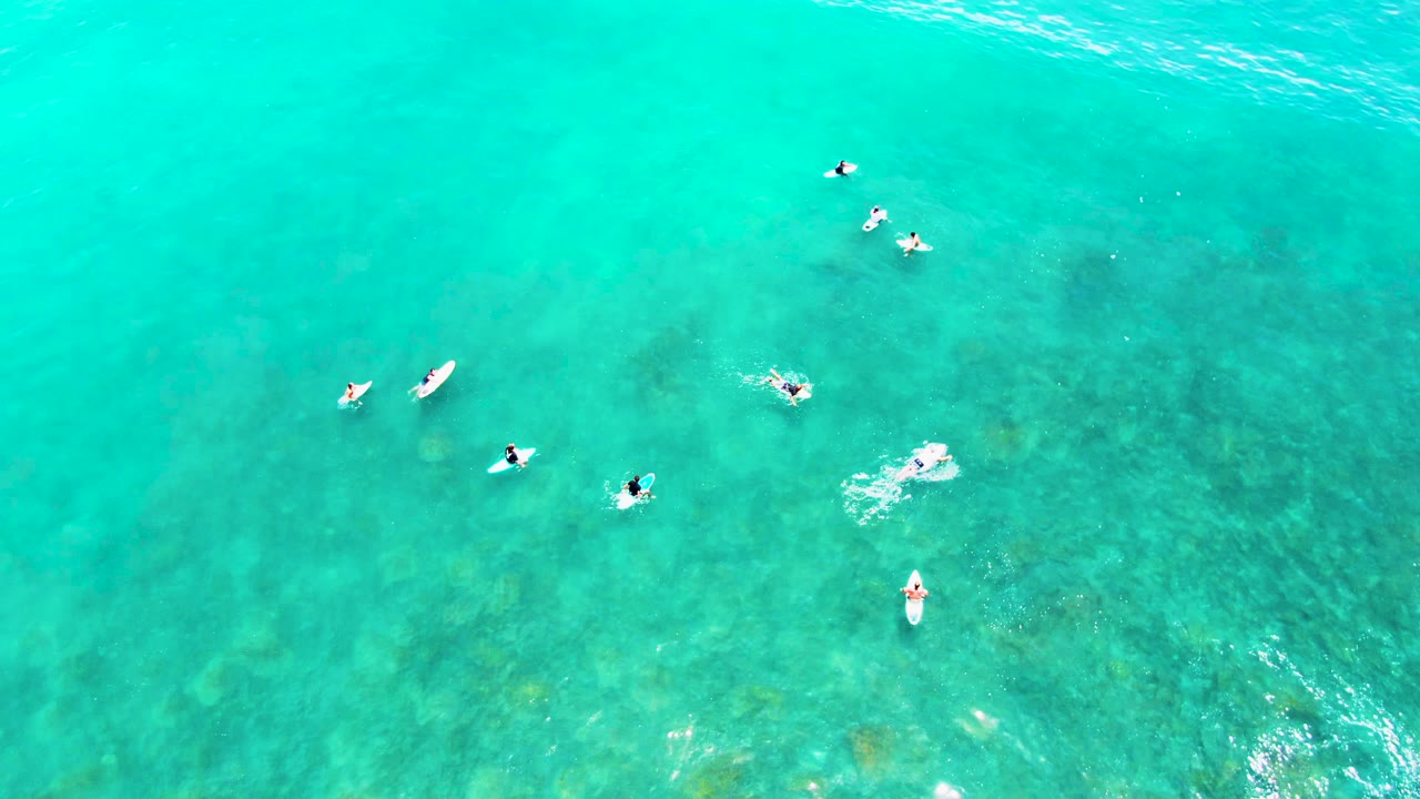 Overhead view at Punta Buzzards, Baja California Sur, showing several swimmers and small boards spaced across clear turquoise water with darker seabed patches below.