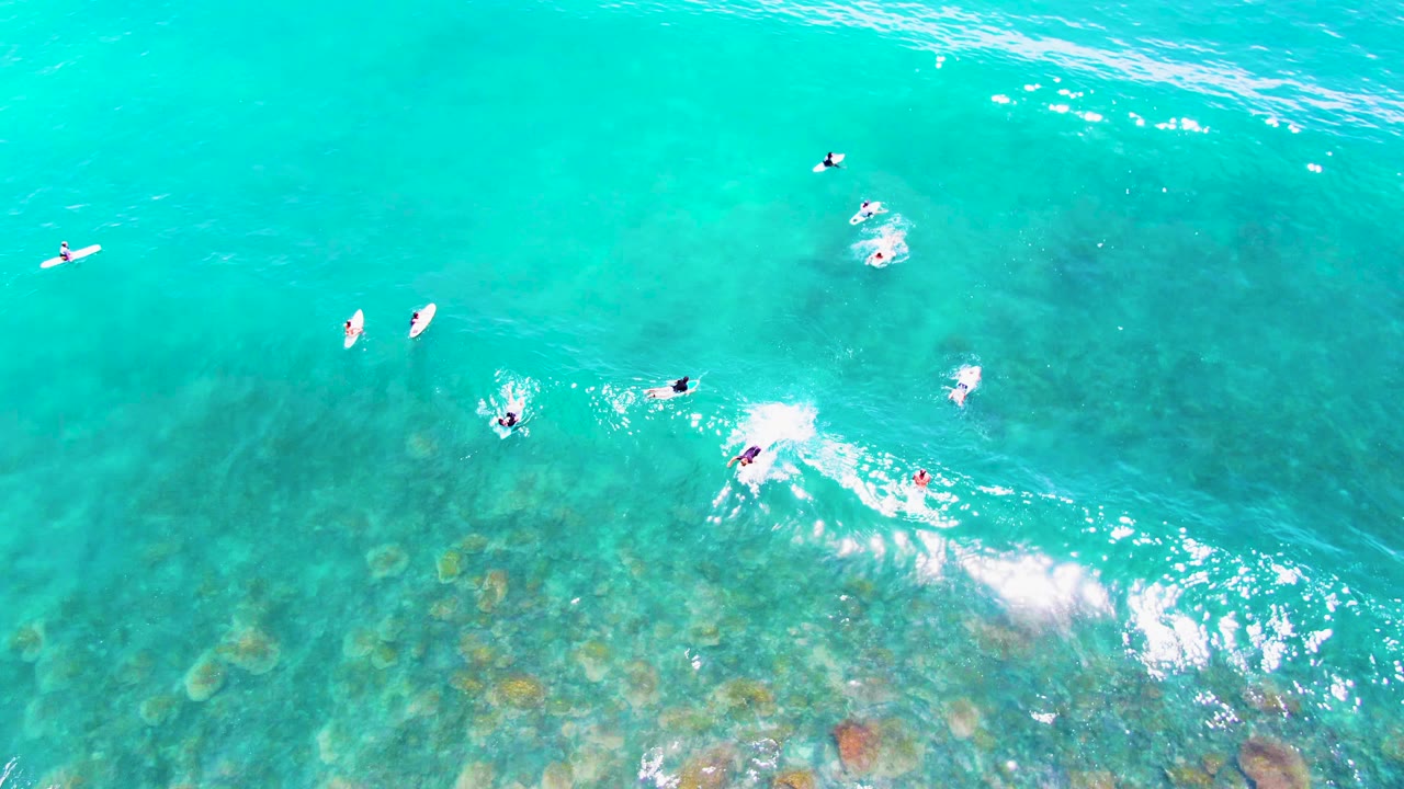 Overhead aerial frame at Punta Buzzards shows scattered surfers on boards in turquoise water, with a whitewater burst and darker ocean-floor patches below.
