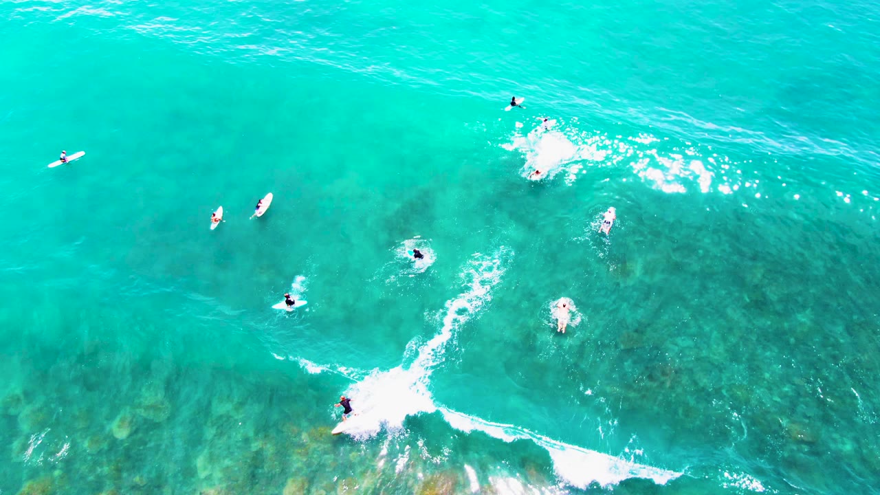 At 0:02, small surfers are spread across the turquoise surface at Punta Buzzards while a bright white breaking wave crosses the lower right of the frame.