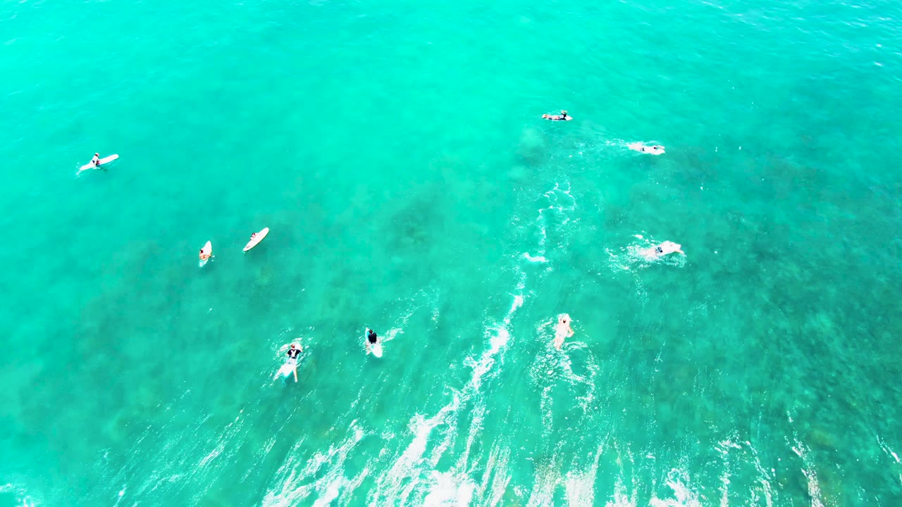 Overhead frame from Punta Buzzards, Baja California Sur showing scattered surfers and swimmers in bright turquoise water with white wash near the lower edge.