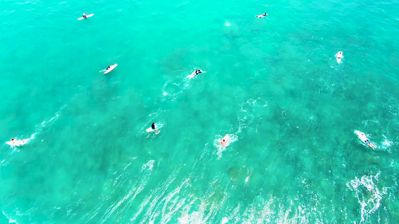 Overhead view at Punta Buzzards with surfers on boards across bright turquoise water, darker ocean-floor areas beneath, and white foam streaking across the lower part of frame.