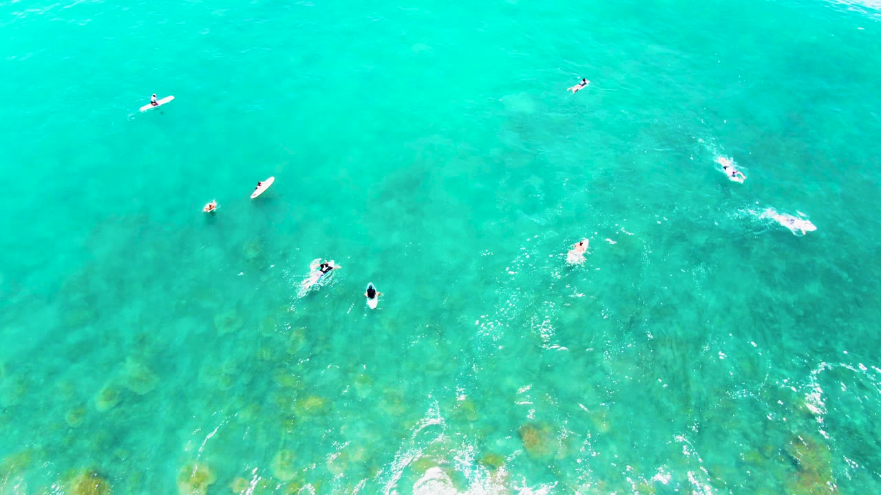 High aerial frame over Punta Buzzards, Baja California Sur, showing scattered surfers on boards in clear turquoise water with darker reef patches and light foam beginning near the lower edge.