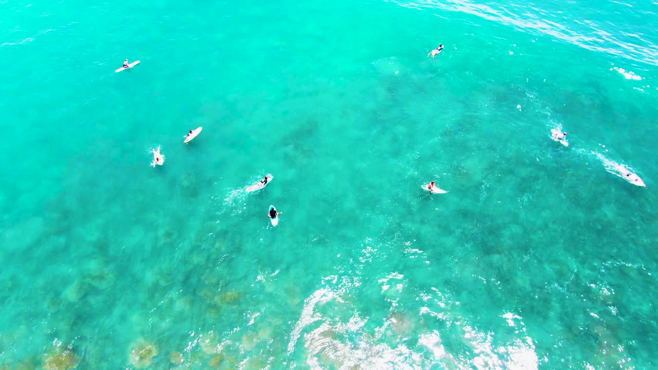 Overhead frame from Punta Buzzards showing several surfers on boards across open turquoise water, with a small paddling wake near center and faint reef color below.