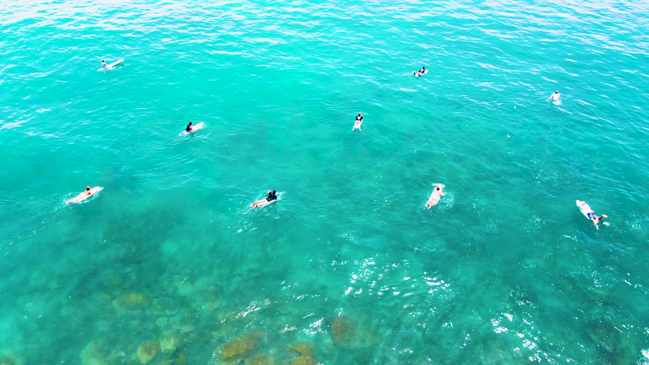 Overhead aerial frame at Buzzards Beach, East Cape, Baja California Sur, with small swimmers and boards spread across clear turquoise water near darker underwater patches.