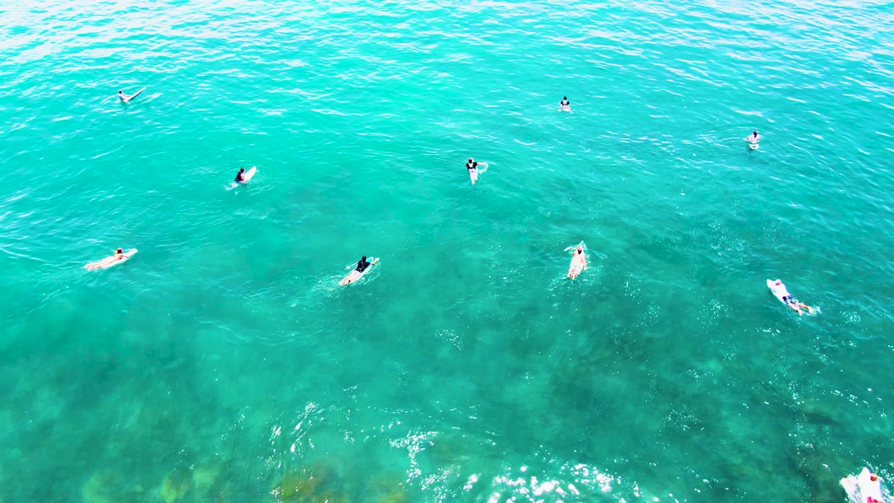 Swimmers and boards float across bright turquoise water from above.