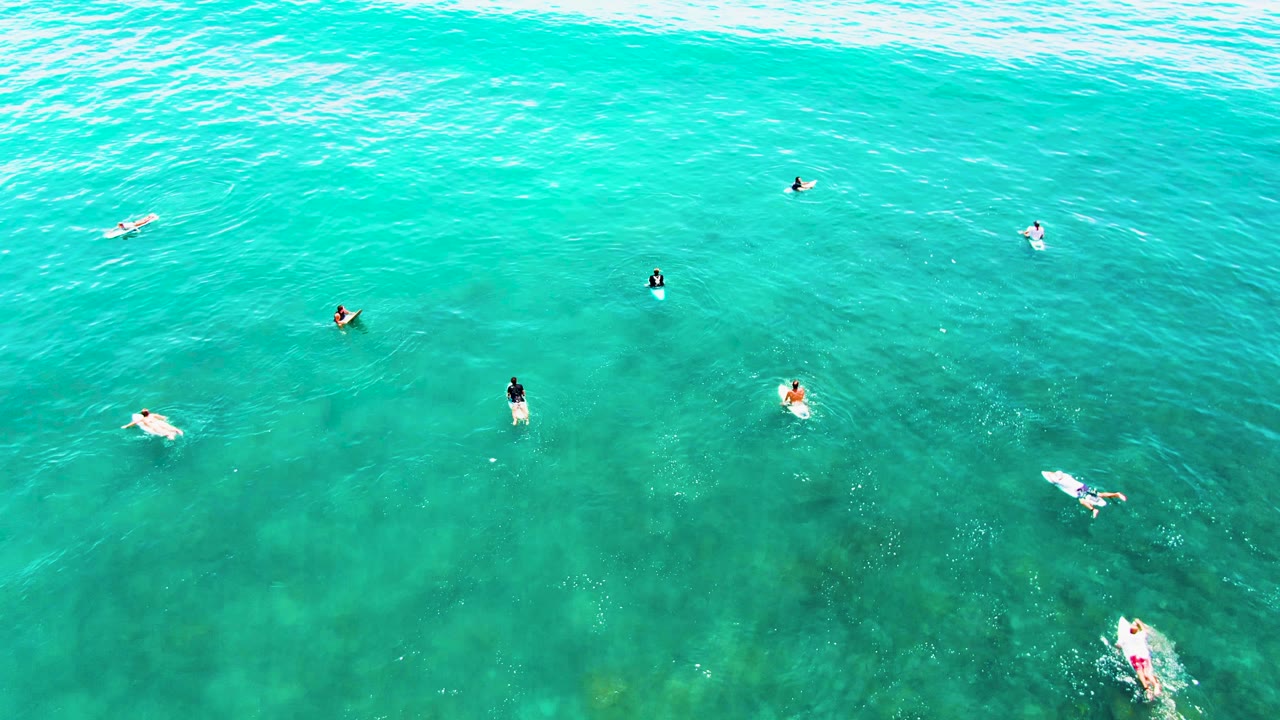 Overhead view at Punta Buzzards, Baja California Sur, showing small swimmers and board riders spread across clear turquoise water as a pale wave band forms near the top of frame.
