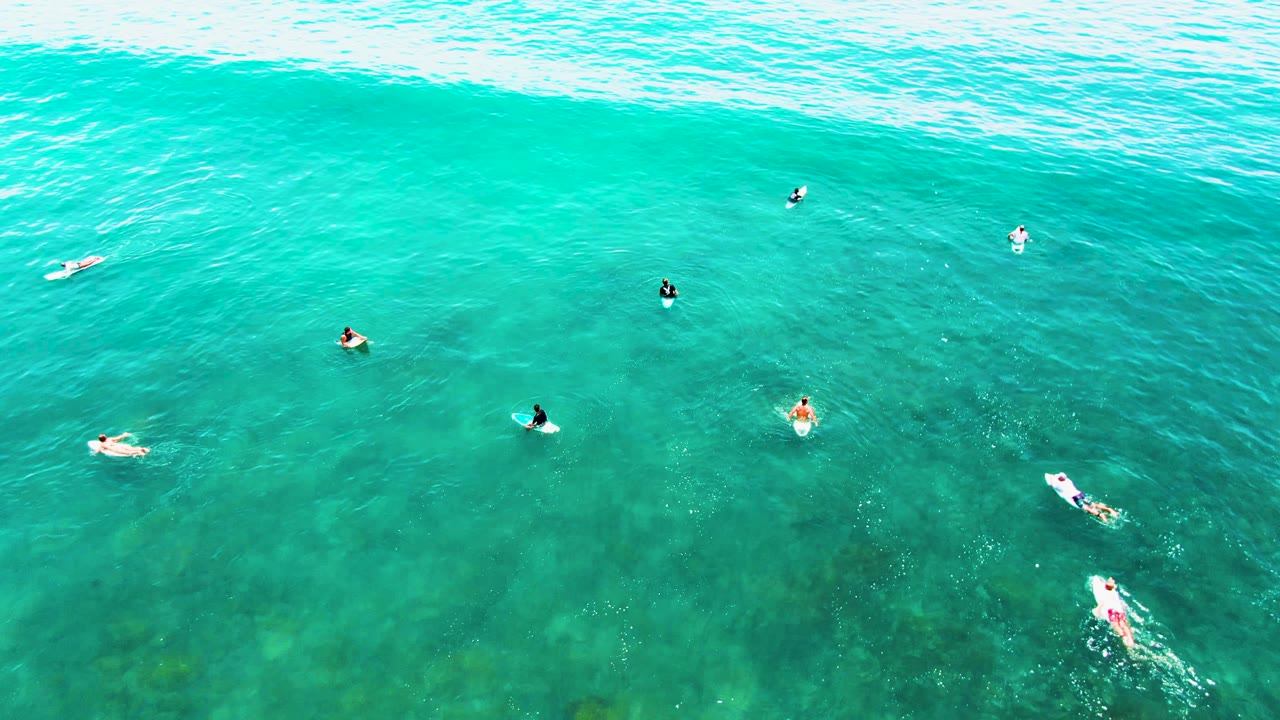 Overhead view of clear ocean at Punta Buzzards with swimmers and board riders separated across the frame and darker green-blue underwater patches visible beneath them.