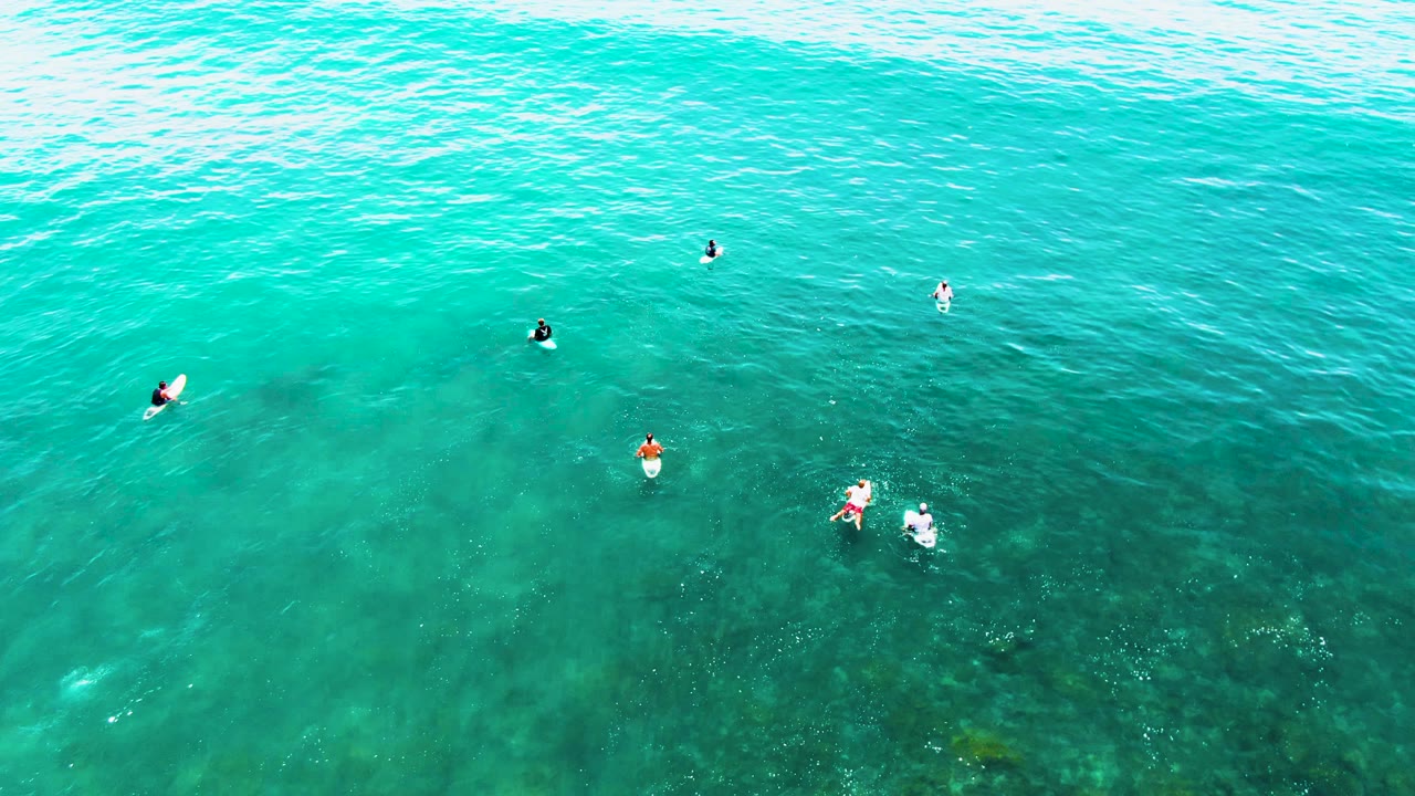 At 0:02, several people remain visible from above in the clear Punta Buzzards ocean, with one pair closer together near the lower right and darker sea floor beneath them.