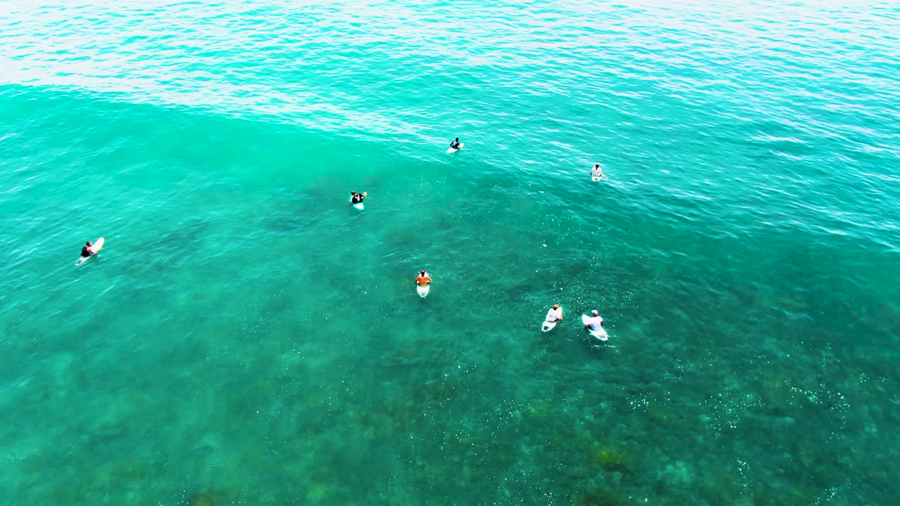 Overhead view at Punta Buzzards, Baja California Sur, with scattered surfers on boards in clear turquoise water and darker seabed patches below.