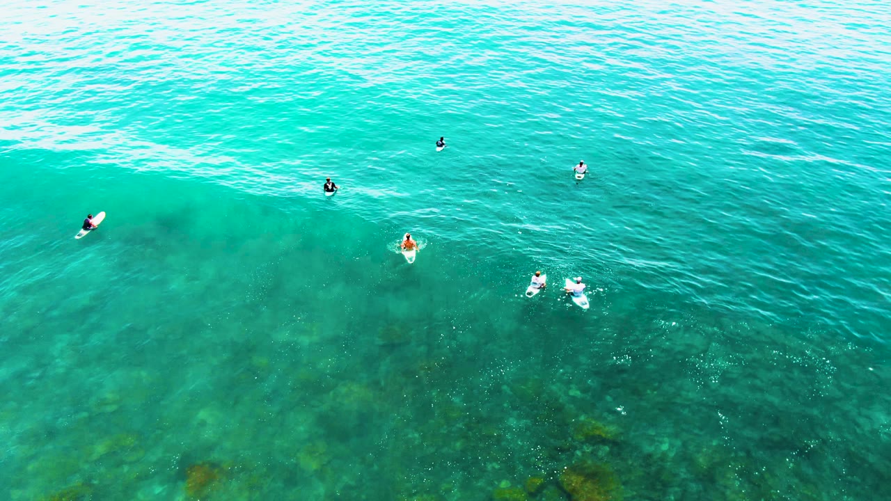 An overhead frame shows several small surfers in Baja California Sur water, with the wave line crossing behind them and darker underwater areas visible below.