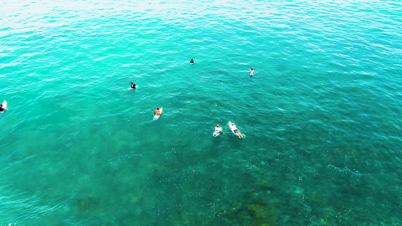 Overhead aerial frame at Punta Buzzards shows several people spread across clear turquoise ocean water with darker underwater patches near the lower half.