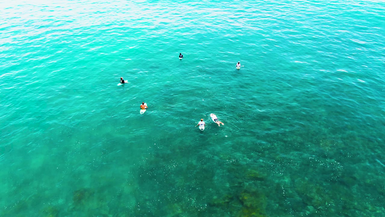 At 0:02, swimmers remain visible across the blue-green water, with subtle wave texture and darker seabed areas showing through the lower right portion.