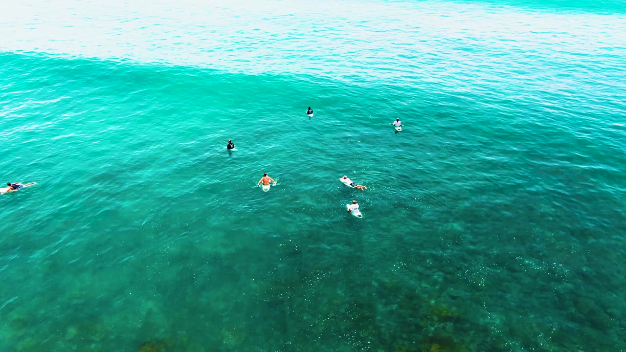 Aerial frame from Punta Buzzards, Baja California Sur, showing scattered surfers and swimmers in clear turquoise water with a pale wave band near the top of the image.