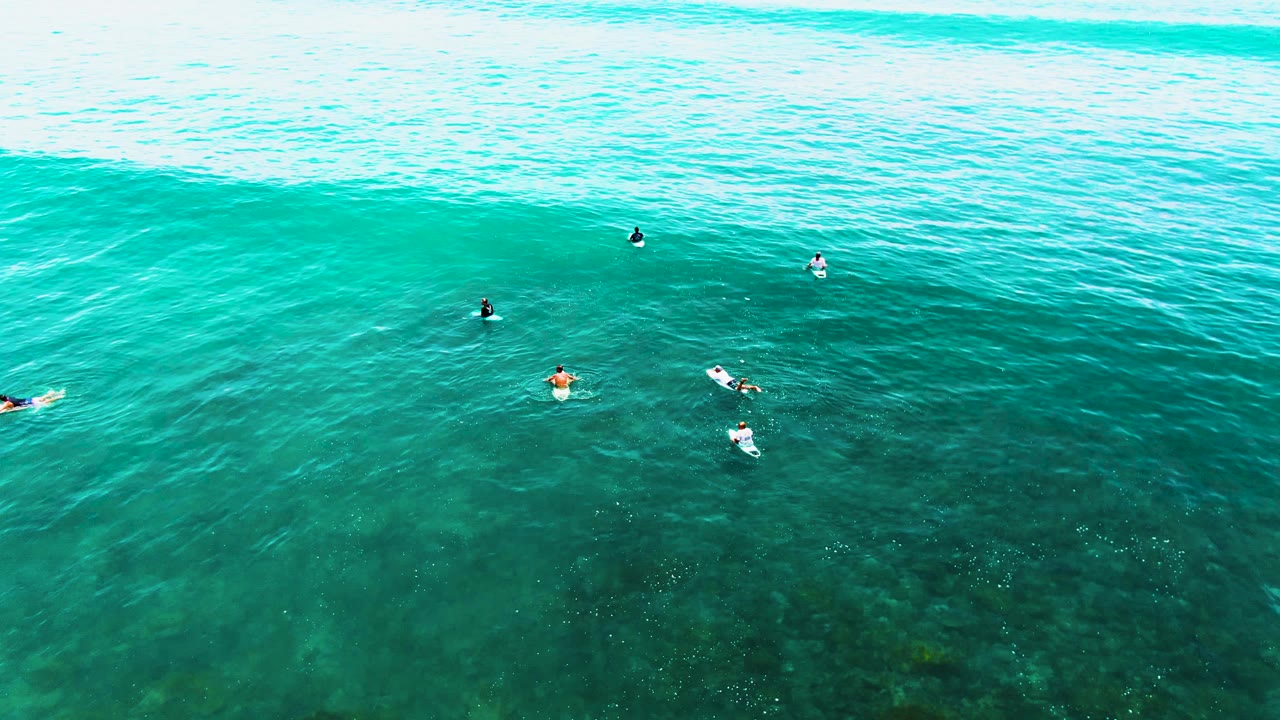 Surfers on boards spaced across clear ocean water below a broad wave line.