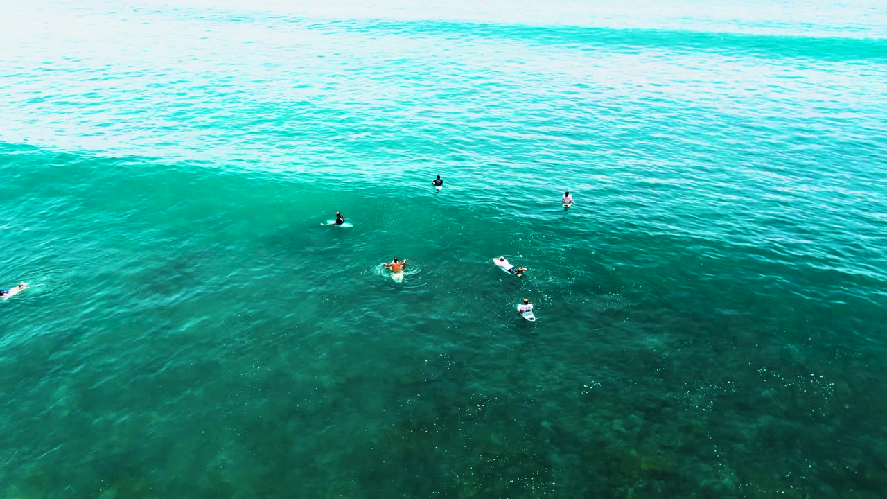 Aerial frame from Punta Buzzards showing surfers and swimmers positioned below a wide green-blue wave face, with dark underwater texture visible beneath them.