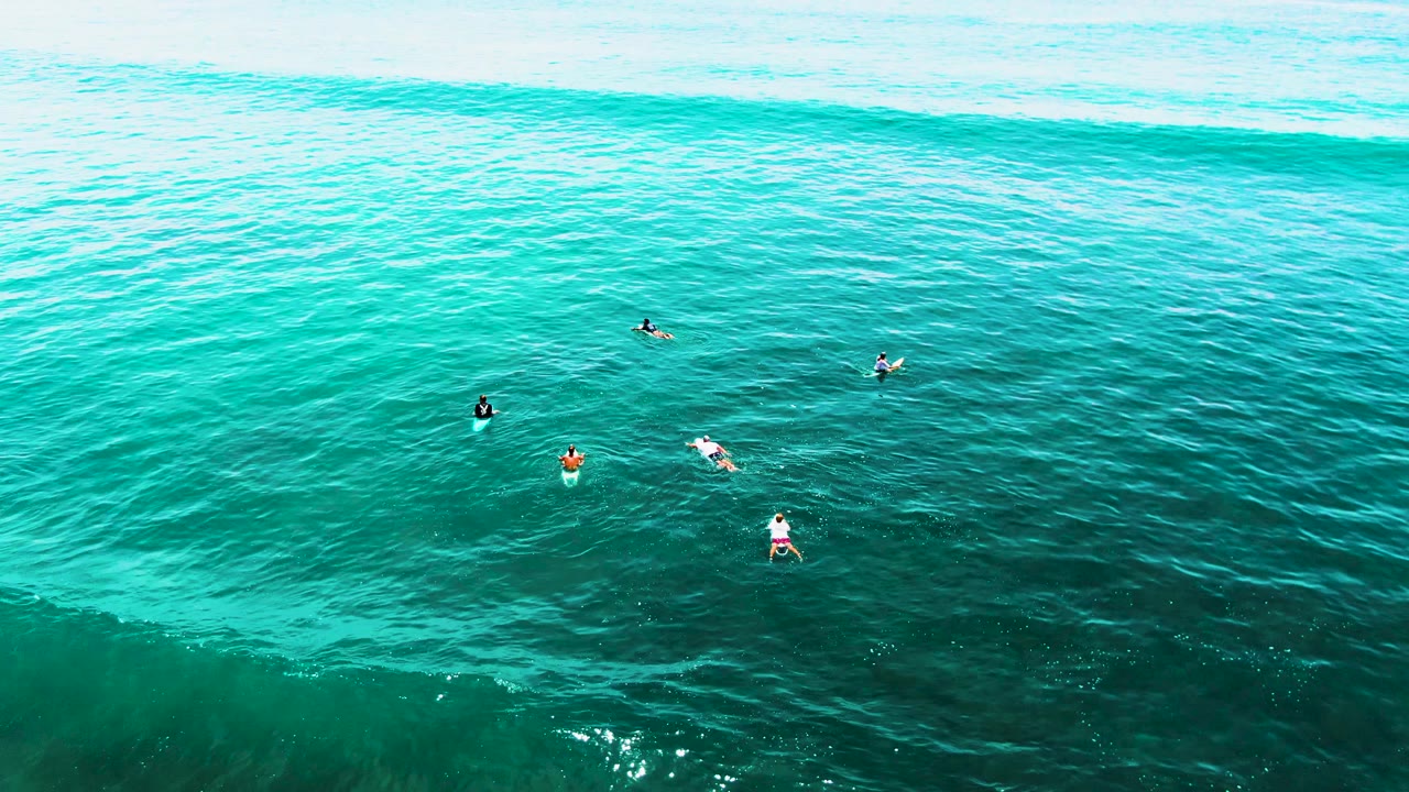 Aerial view at Punta Buzzards, Baja California Sur, showing several surfers spaced across turquoise water as a broad wave line forms near the top of the frame.
