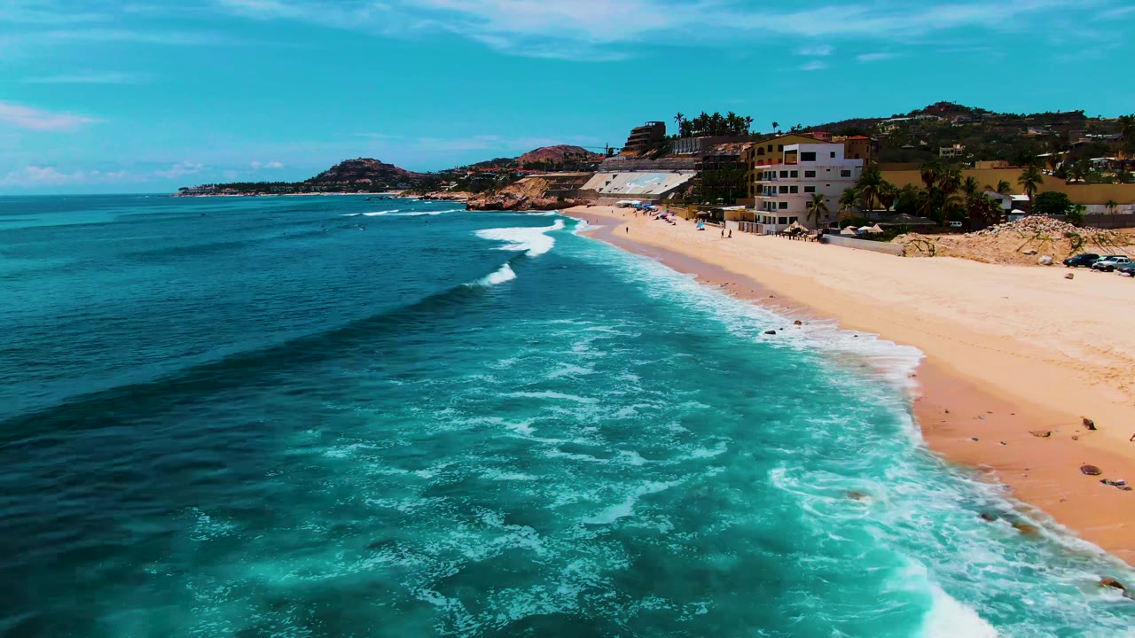 Aerial view at Punta Buzzards, Baja California Sur, with deep blue ocean, turquoise surf, a sandy beach, hillside buildings, and no visible people.