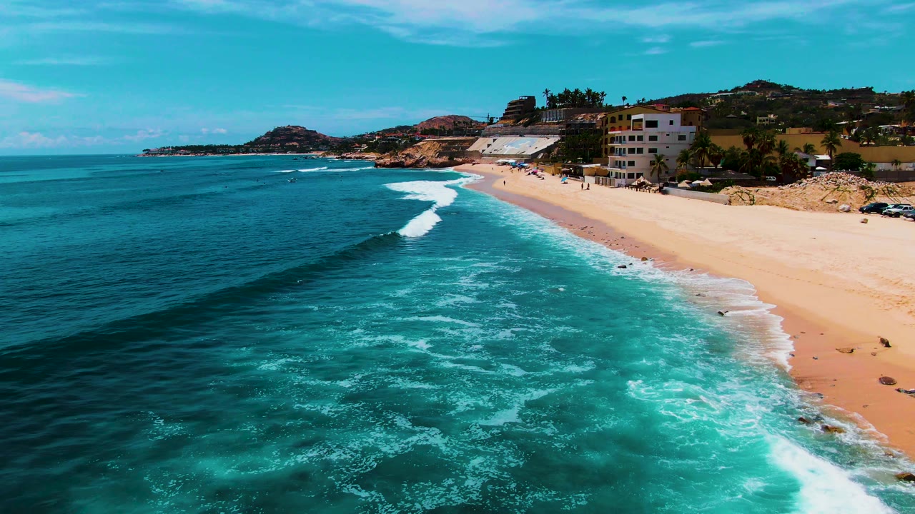 At 0:01, the Punta Buzzards shoreline shows a longer white foam band curling onto sand below hillside buildings, with no visible people.