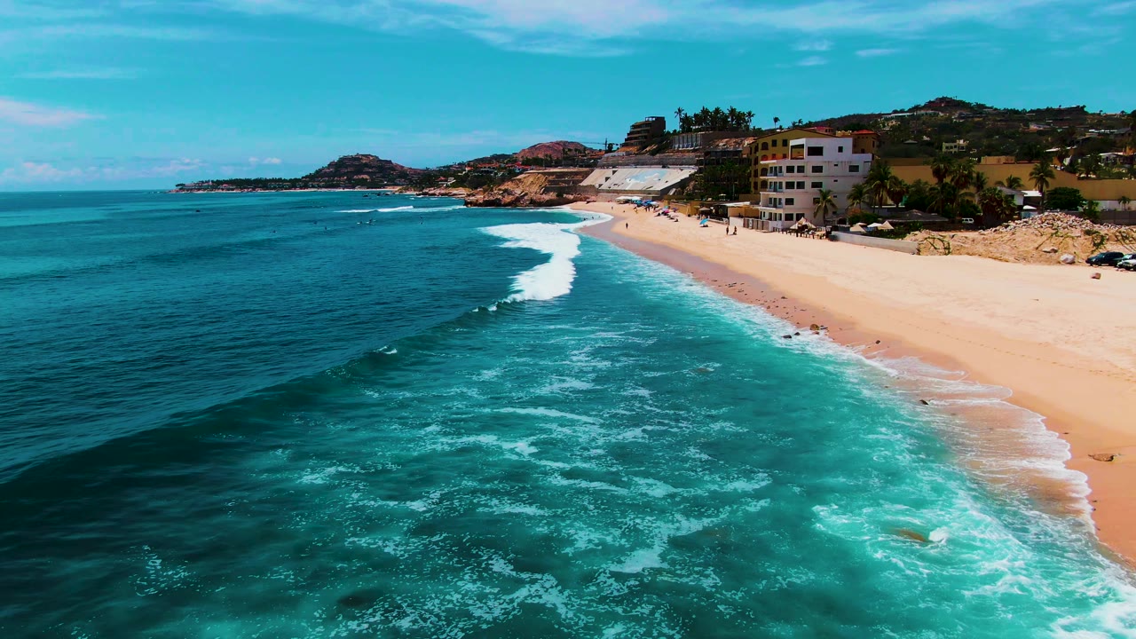 At 0:02, white foam spreads across turquoise water beside the sandy Punta Buzzards beach, with hillside buildings and no visible people.