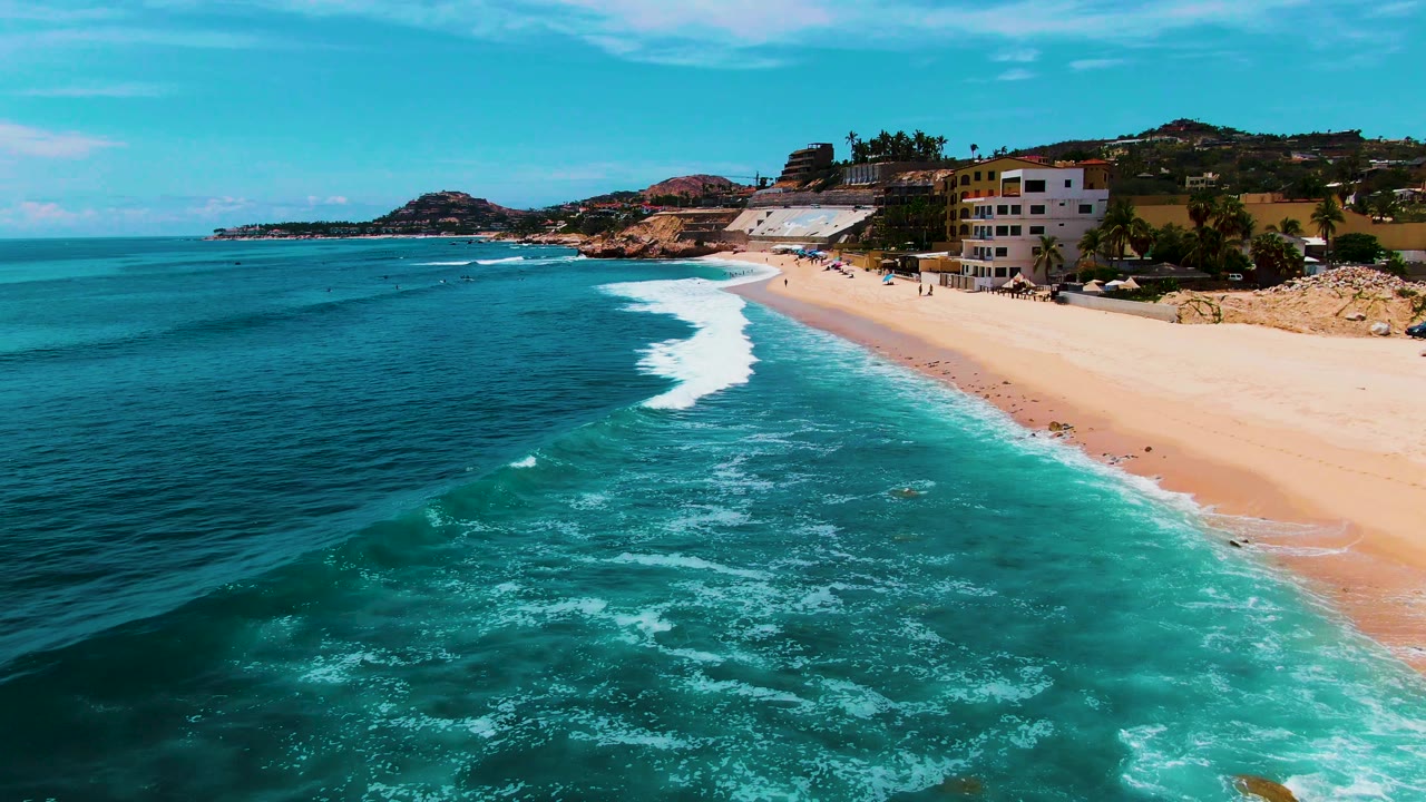 Diagonal shoreline with dark blue ocean, turquoise water, and waves reaching pale sand.