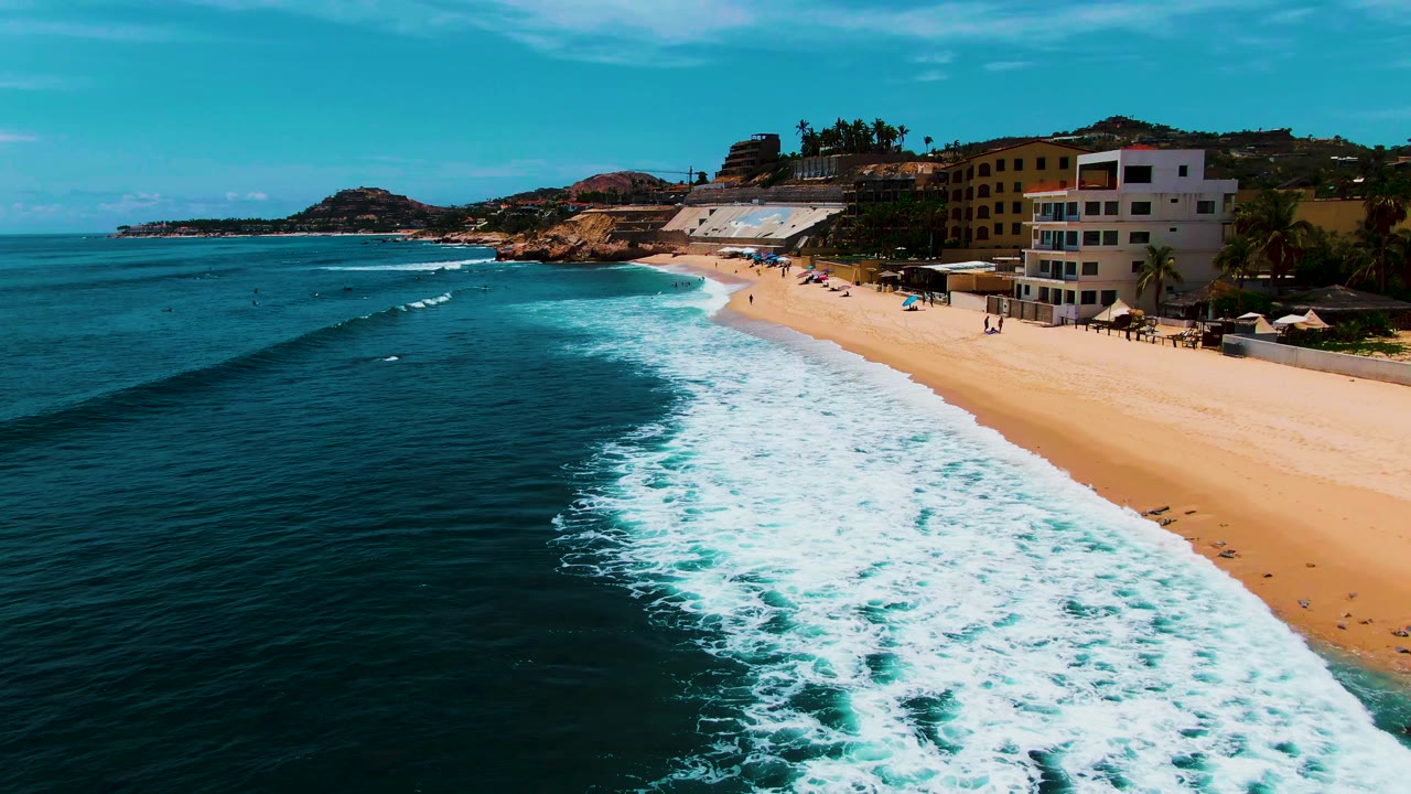 Aerial frame of Punta Buzzards, Baja California Sur, showing turquoise ocean and white surf beside a sandy beach with small visible beachgoers and buildings along the shore.