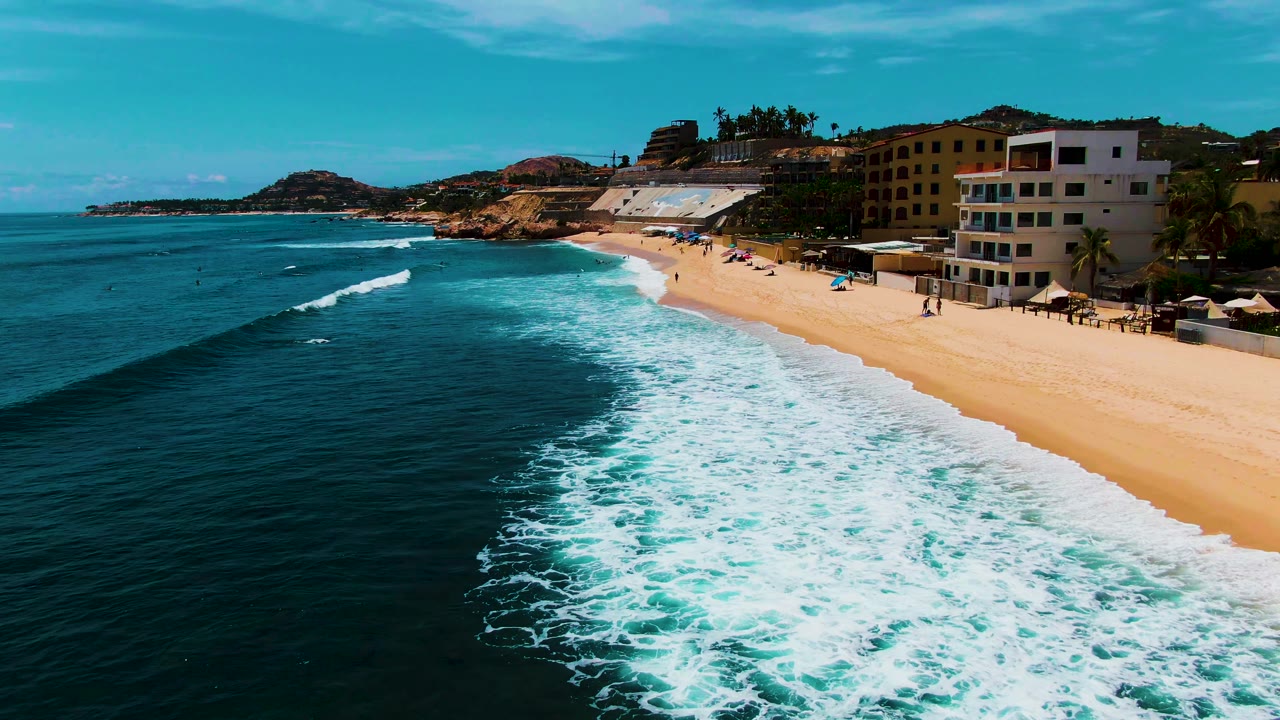 Wide aerial coastline with foamy surf reaching a sandy beach and buildings behind it.