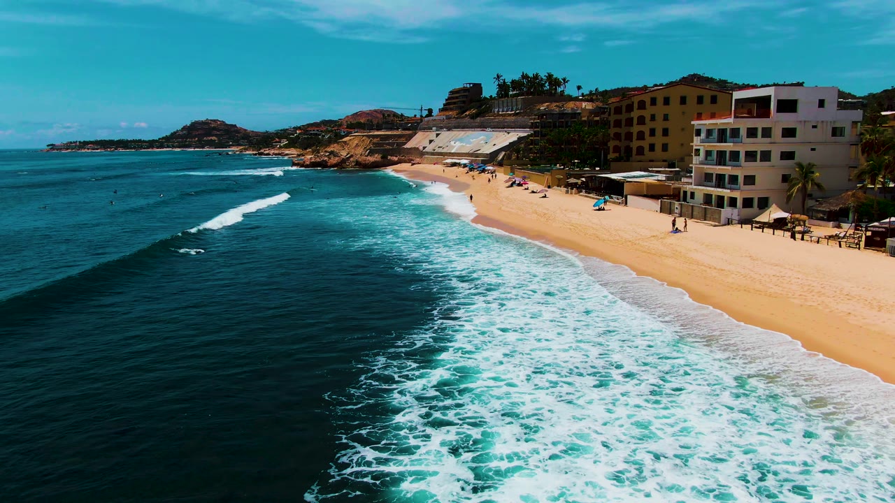 Punta Buzzards beach from above, with the surf line curving through turquoise water, beachgoers spaced along the sand, and a white multi-story building near the right edge.