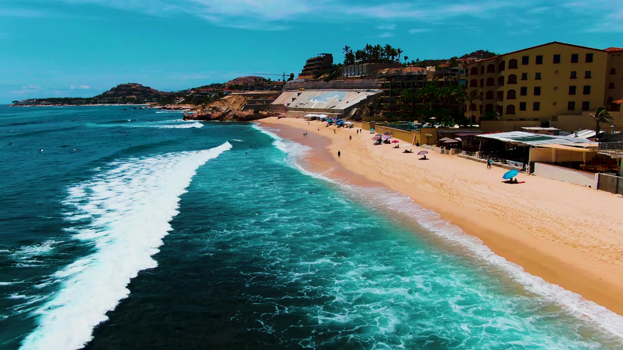Aerial view of Buzzards Beach in Baja California Sur with turquoise surf breaking toward a sandy shore, beach umbrellas, small beachgoers, coastal buildings, palms, and a rocky headland in the distance.