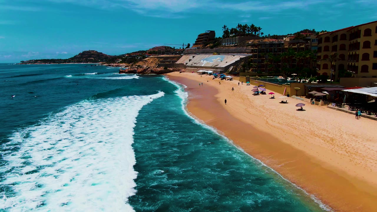 Aerial coastline view at Buzzards Beach with a curved white wave front, turquoise water, open sand, beach umbrellas, small people near the shore, buildings on the right, and rocky coast in the background.
