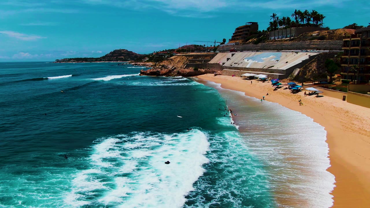 Aerial view at Punta Buzzards in Baja California Sur with white surf rolling toward a sandy beach, small swimmers in the water, umbrellas on shore, and rocky coastline beyond.