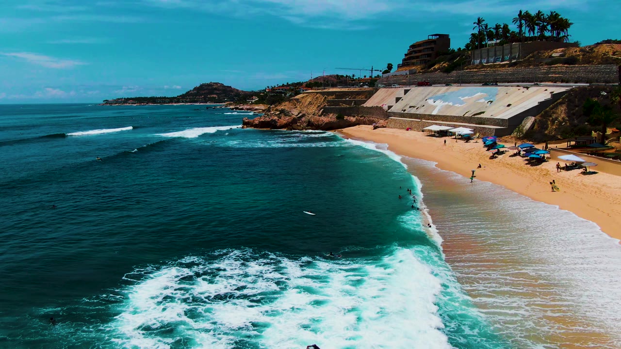Foamy surf spreads across turquoise water near a sandy beach with people in the shallows.