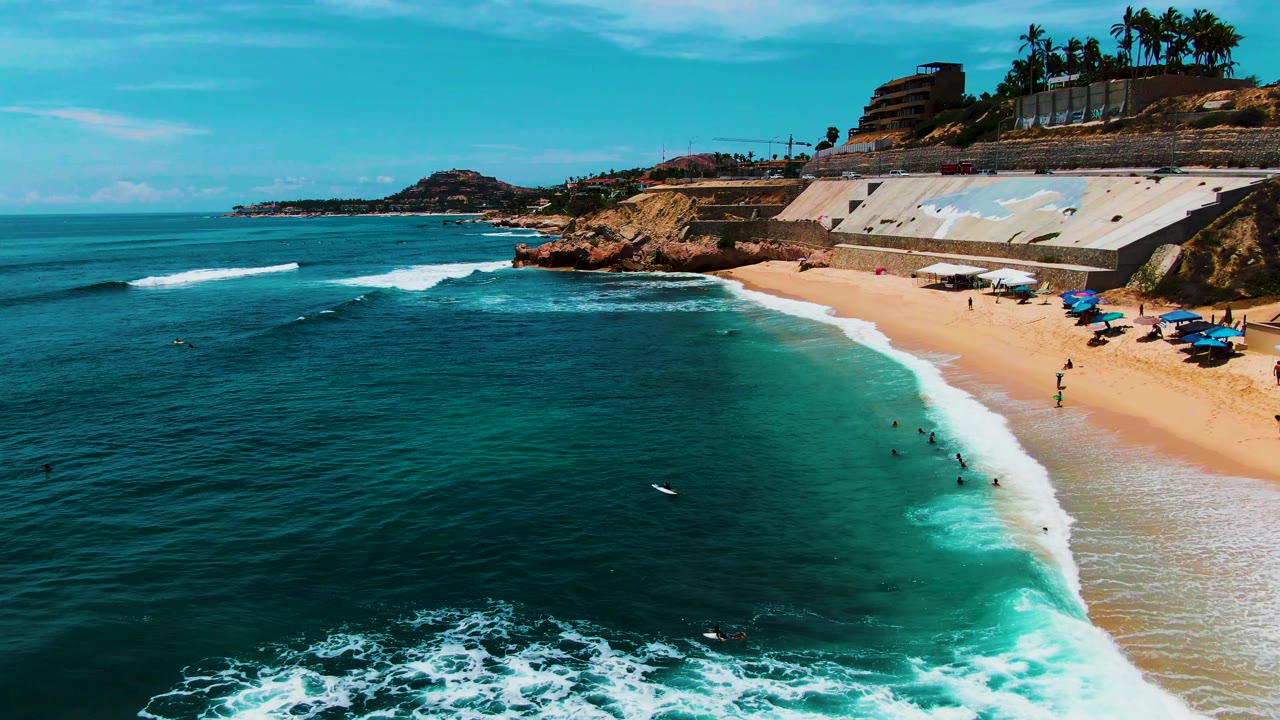 At 0:02, the aerial view holds over Punta Buzzards with swimmers spaced along the foamy shorebreak, a rocky point to the left, and seawalls and palms above the sand.