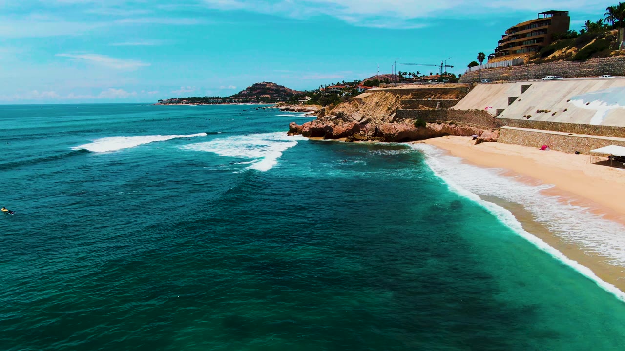 Aerial view at Buzzards Beach in Baja California Sur with deep blue ocean, a narrow white foam line, sandy shore, rocky cliffs, cliffside buildings, and no visible people.