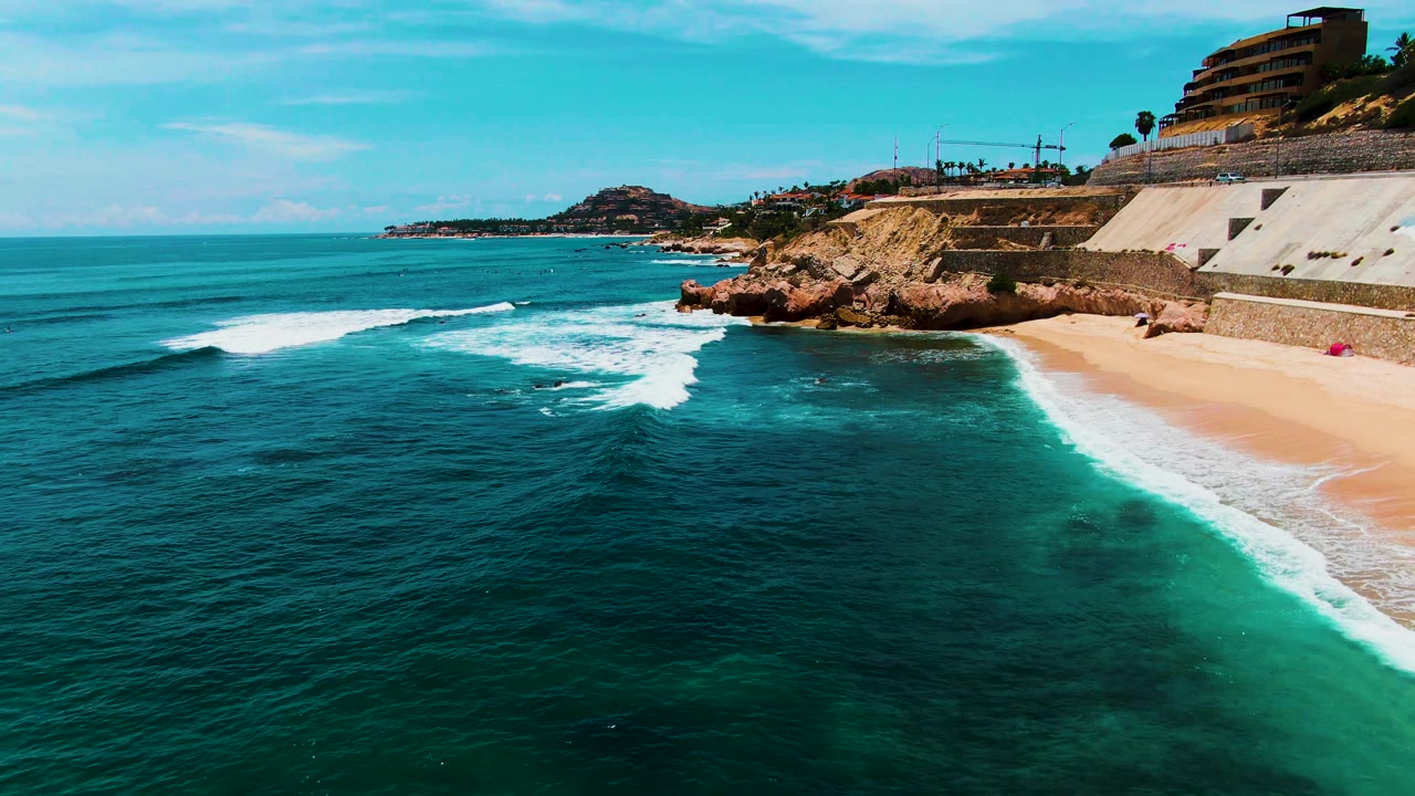 At Buzzards Beach in Baja California Sur, a broader band of white surf moves over turquoise water toward the sand, with rocky cliffs and buildings along the upper right, and no visible people.