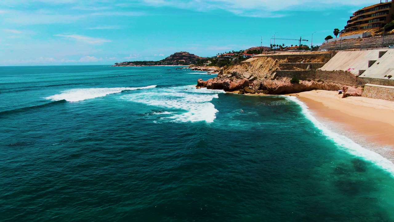 A breaking wave spreads white foam across the nearshore water at Buzzards Beach in Baja California Sur, with sand at right, rocky cliffs behind it, and no visible people.