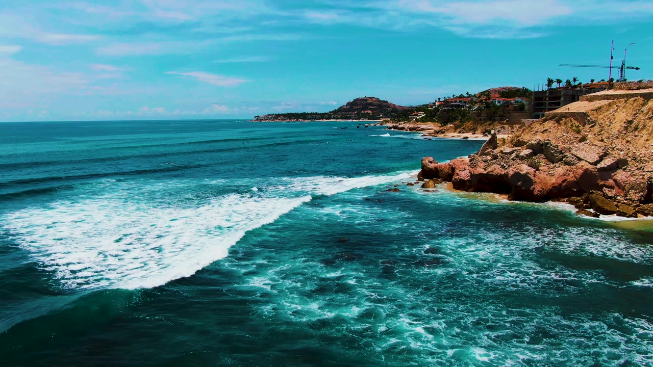 Wide aerial view of turquoise surf breaking beside rocky coast and sandy beach.