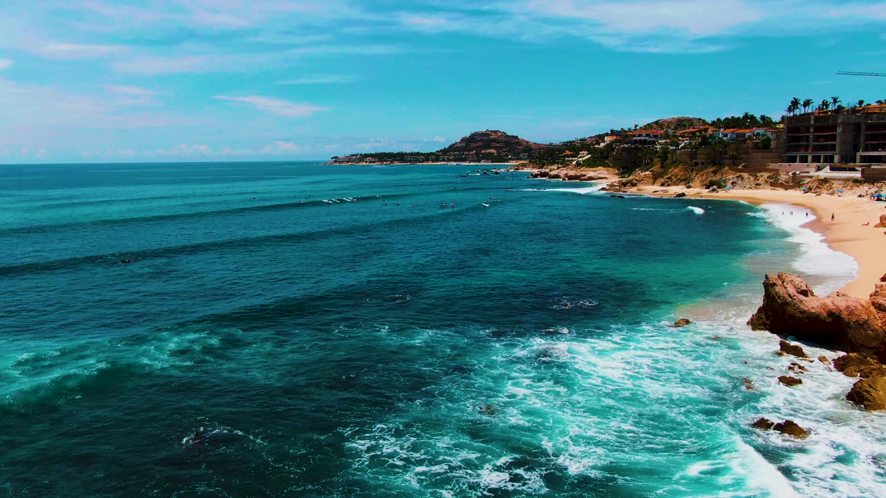 Aerial view at Punta Buzzards, Baja California Sur, with turquoise ocean in the foreground, white surf on a sandy beach, dark rocks near the lower edge, and hillside buildings in the distance.