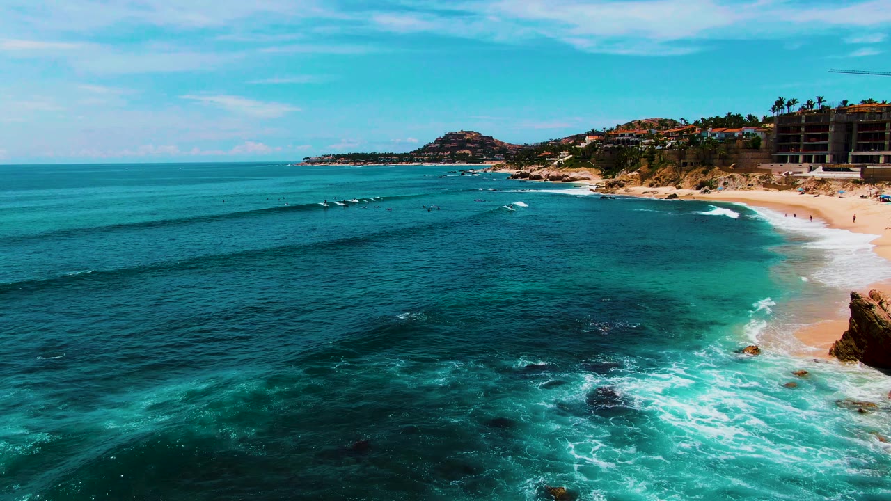 Punta Buzzards aerial frame with darker reef-like water patches in the foreground, white foam near the lower right rocks, and a sandy beach backed by hillside structures.