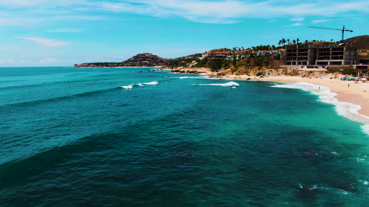 At Punta Buzzards in Baja California Sur, turquoise ocean stretches toward a sandy beach with hillside buildings and a construction crane in the distance.