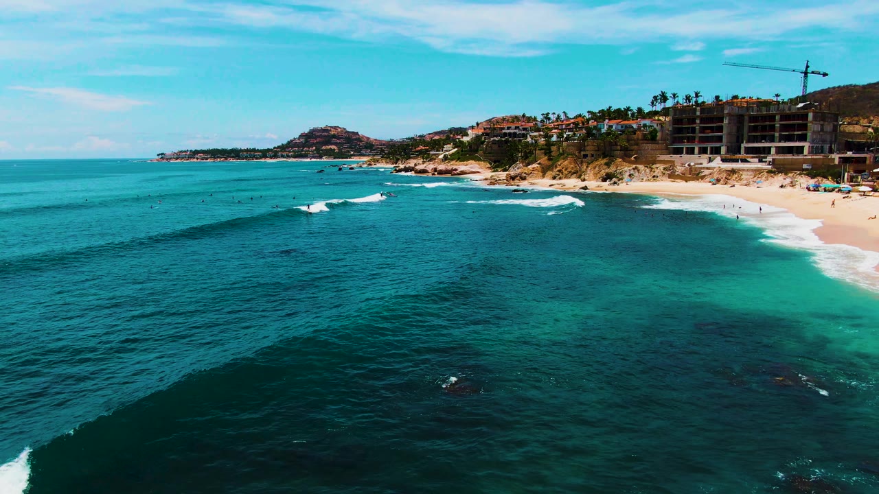 Wide aerial beach view with waves approaching shore and construction on the hillside.