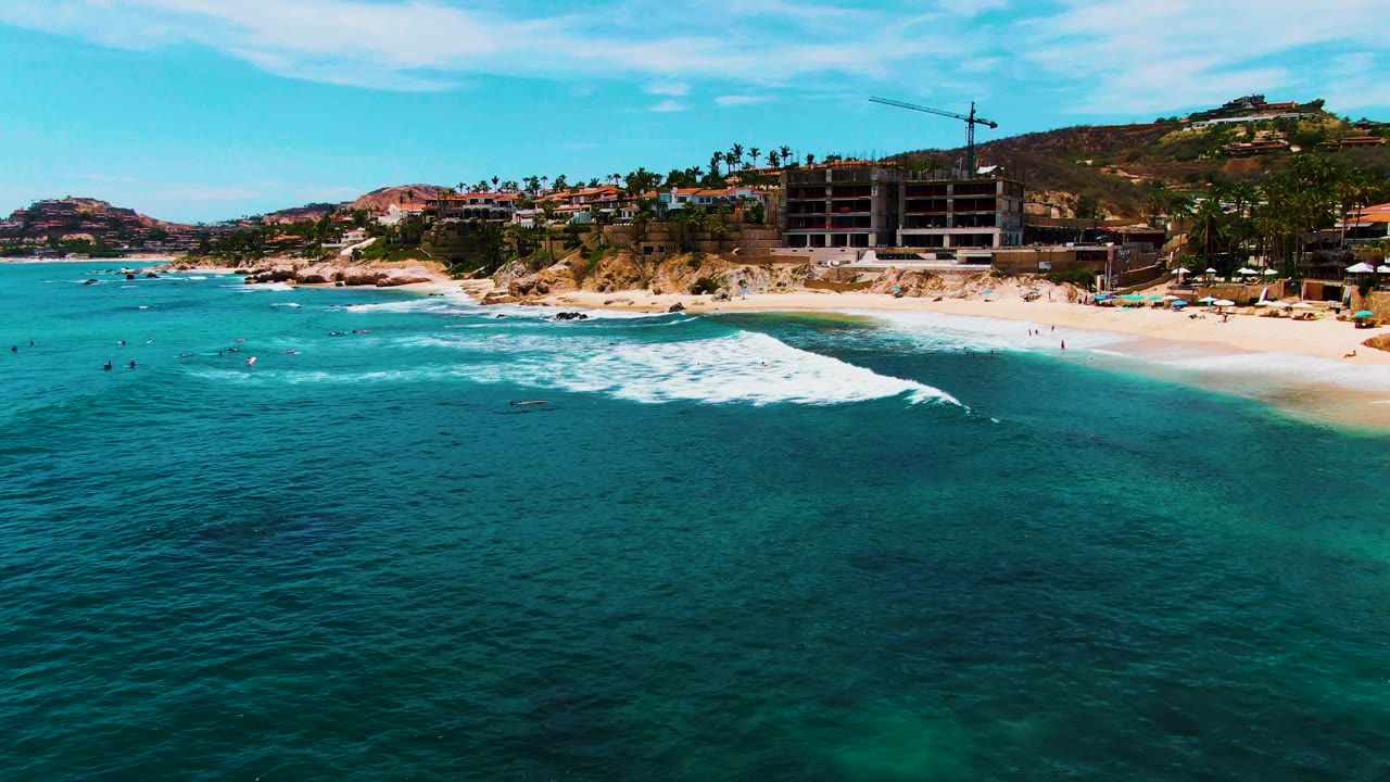 Aerial view of Buzzards Beach in Baja California Sur, Mexico, with turquoise water in front, a white wave nearing the sand, small swimmers offshore, and hillside buildings behind the beach.