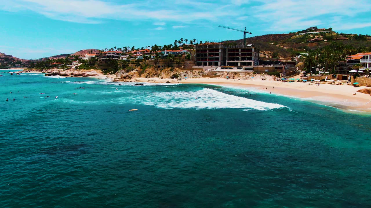At 0:02, the aerial view holds on Buzzards Beach as swimmers dot the turquoise water, a foamy wave reaches the sand, and hillside buildings and a crane remain visible above the shoreline.