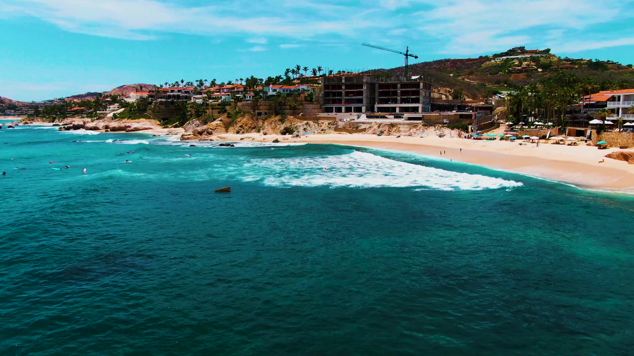 Foam spreads across turquoise water in front of a sandy beach and hillside buildings.